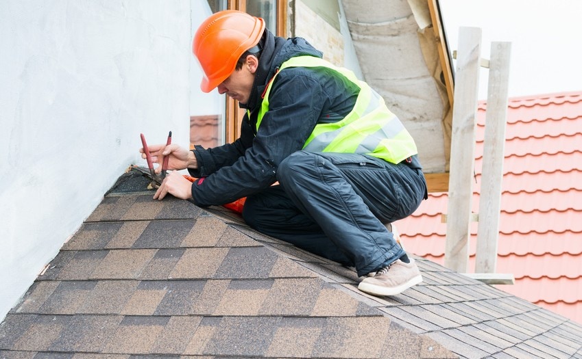 A roofer, wearing an orange hard hat and a high-visibility vest, is kneeling on a gabled section of a roof. He is using a pair of pliers or snips to work on the edge of the roof, which is covered in brown shingles. The roofer is working on the edge where the roof meets a light-colored, stucco-like wall. Another roof section with red tile is visible in the background.