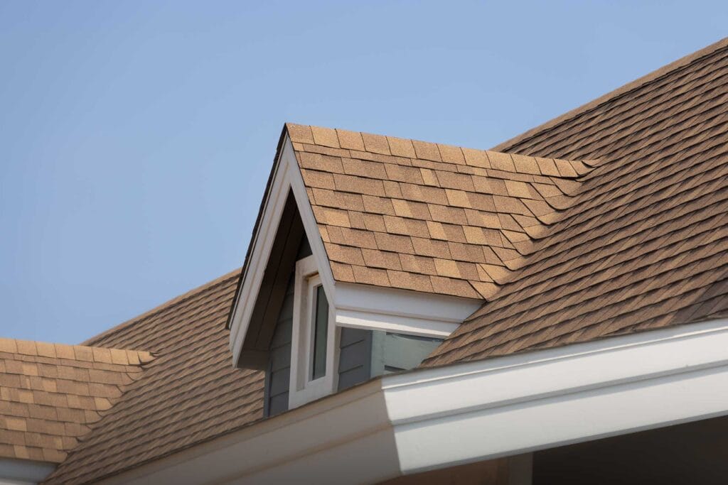A close-up of a house roof with brown asphalt shingles showcases the craftsmanship you'd expect from a top roofer in New Castle County. The roof features a dormer with a small gable and white trim, set against a clear blue sky, highlighting the skill involved in roof repair.