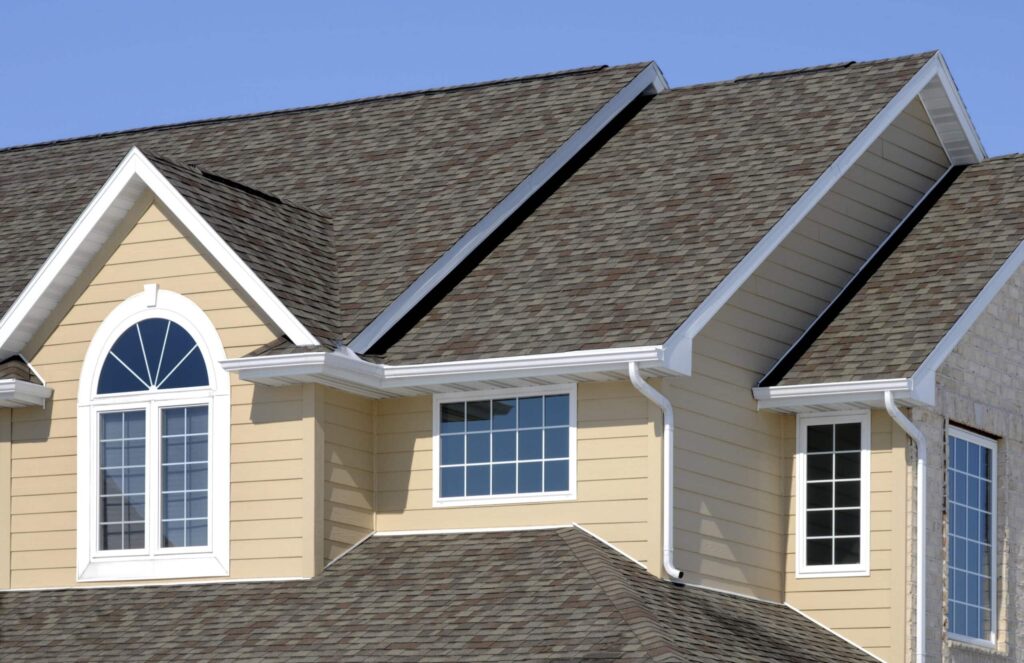 A close-up of the upper half of a two-story house with a brown shingled roof. The house has light yellow horizontal siding with white trim, gutters, and downspouts. Several windows are visible, including a large arched window with white muntin grids on the left. The sky is a clear blue.