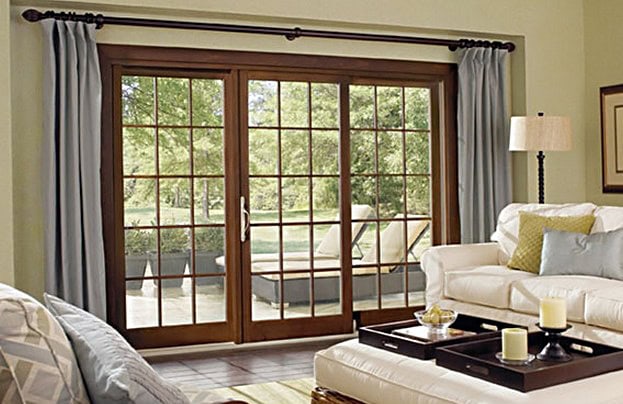 A cozy living room with a white couch, a floor lamp, and a coffee table adorned with candles. Large sliding glass doors open to a patio with outdoor furniture, revealing a lush garden view. Light gray curtains frame the doors; consider roof repair in New Castle County, DE for peace of mind.