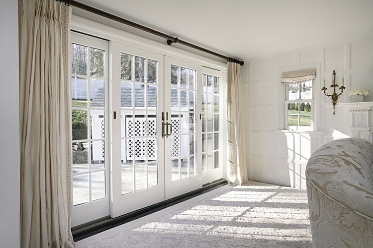 A bright living room with large glass French doors leading outside. Cream curtains frame the doors, and sunlight casts shadows on the light carpet. A patterned sofa and a wall sconce are visible in the corner, all under a roof maintained by expert roofers in New Castle County, DE.