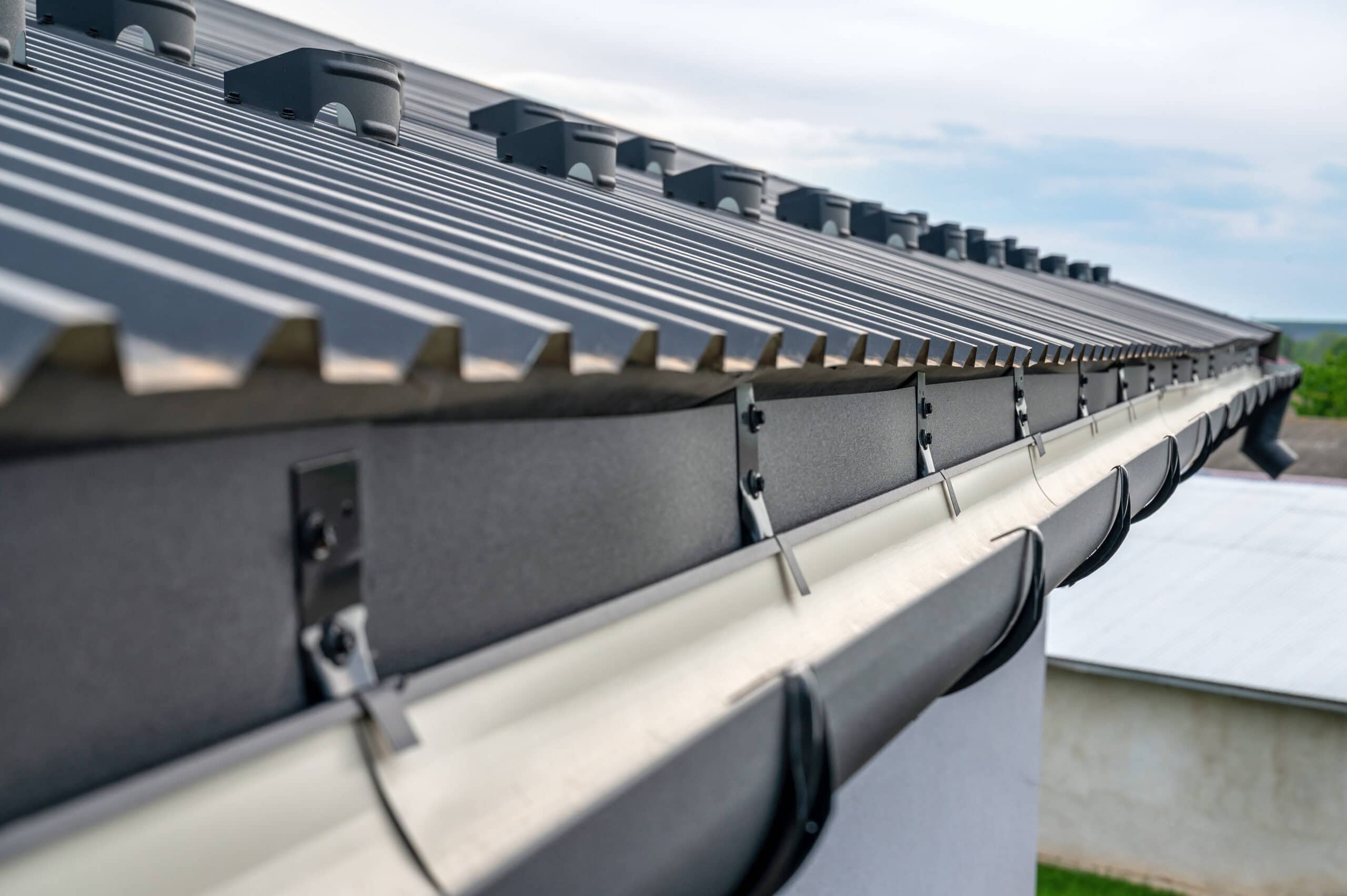 Close-up view of a metal roof in New Castle County, DE, with solar panel mounts along the edge. Below the roof, a white gutter system is attached, curving along the side of a building. The sky is partly cloudy in the background. Perfect for those considering roof repair.