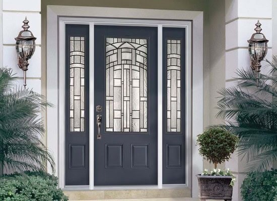 A dark gray front door with decorative glass panels featuring geometric patterns stands proudly, framed by two tall matching side panels. Two ornate wall-mounted lanterns and potted plants complete the elegant entrance, highlighting the craftsmanship found in homes throughout New Castle County, DE.