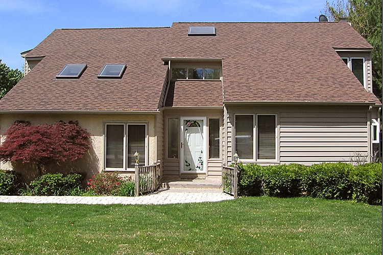 A suburban house with a brown, sloped roof in need of expert roofer services in New Castle County. It features skylights, light-colored siding, a decorative front door, and a path winding through a well-kept lawn and bushes, with a small red tree on the left.