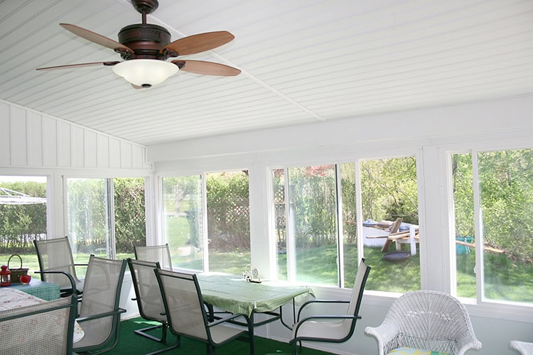 Bright sunroom with white walls and ceiling, featuring a roofer-approved ceiling fan with wooden blades. Large windows offer a view of the lush New Castle County greenery. The room is furnished with a green tablecloth on the table and several wicker and metal chairs.