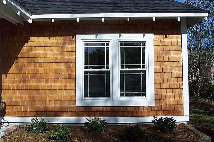 A house exterior in DE showcases brown wooden shingle siding and white-framed double windows. Small shrubs nestle beneath the windows, with trees providing a serene backdrop. A skilled roofer in New Castle County, DE, has ensured the roof complements this cozy home perfectly.