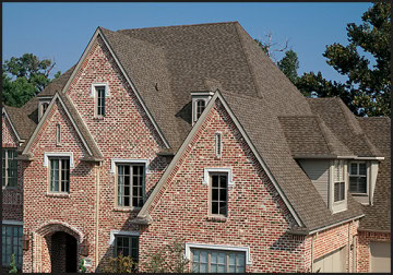 A large brick house with multiple steep gables and gray asphalt shingles recently underwent roof repair in New Castle County. The symmetrically aligned windows on the front facade complement the surrounding greenery and trees beautifully.
