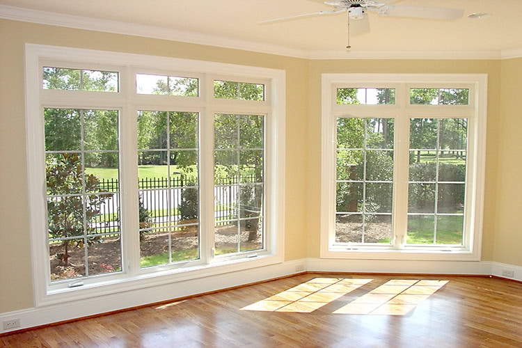 A sunlit room with three large, white-trimmed windows presents a beautiful view of a fenced garden and trees. The light beige walls complement the wooden floor, while a newly installed ceiling fan by DE's finest roofer in New Castle County adds charm from above.