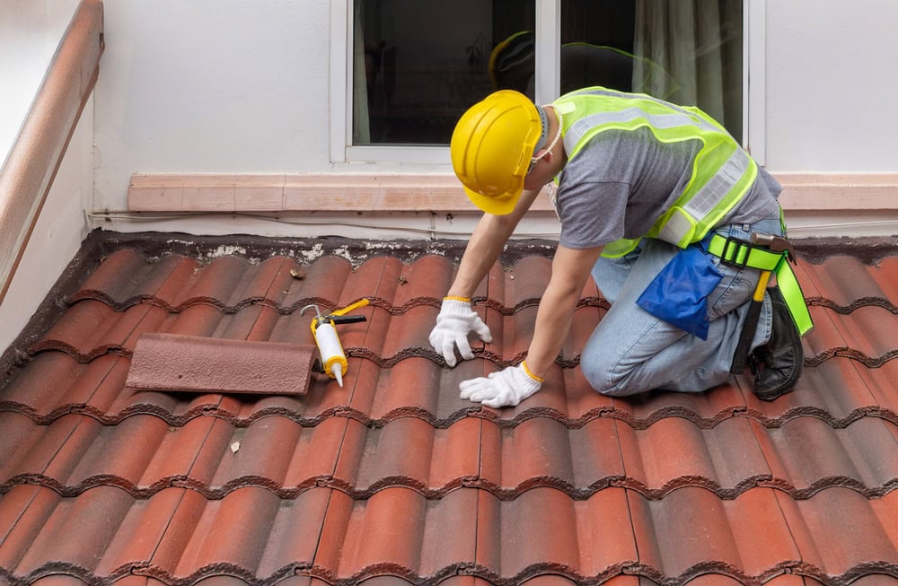 A skilled roofer in a yellow hard hat and safety vest kneels on a red tiled roof in New Castle County, DE, diligently performing roof repair. Equipped with white gloves and tools like a caulking gun nearby, he works with precision as a window frames the background.