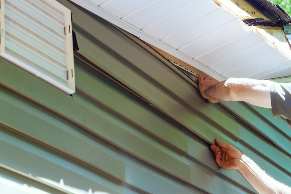 A skilled roofer in New Castle County meticulously installs green vinyl siding, expertly fitting panels under the existing layers below a vent and roofline. In bright daylight, shadows dance across the walls and siding, enhancing the home's fresh appearance.