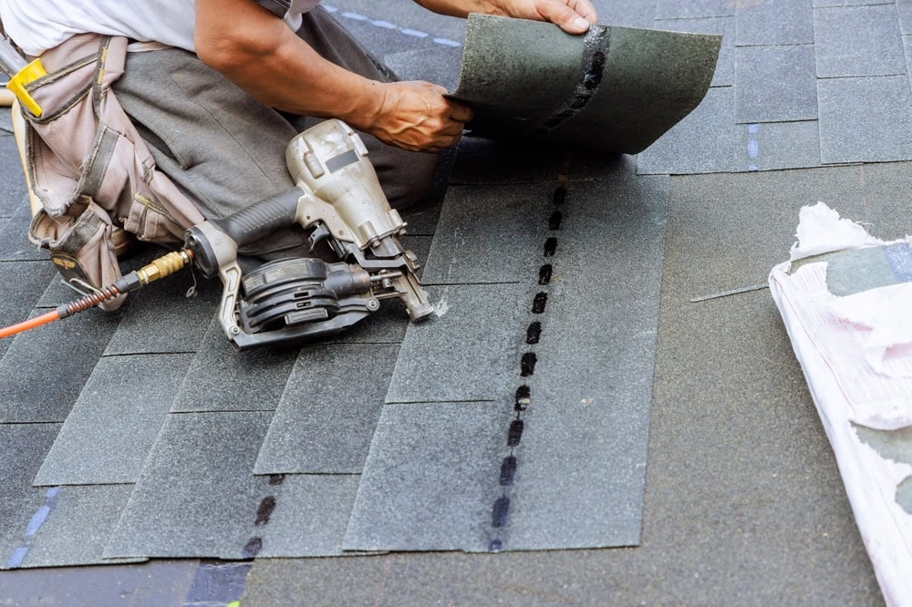A roofer in New Castle County, DE is installing asphalt shingles on a roof. They skillfully hold a shingle in one hand and use a nail gun with the other. With a tool belt strapped on, they work efficiently to complete the roof repair, leaving it partially covered with dark shingles.