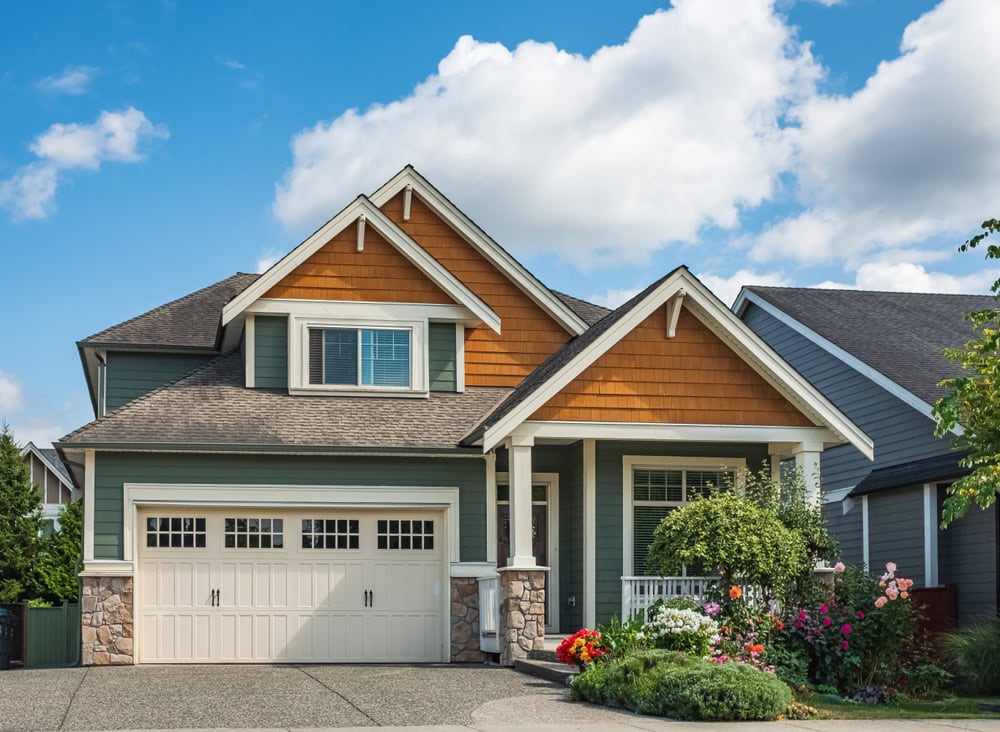 A modern suburban house with green and orange siding, a double garage, white trim, and a covered porch. The front yard features blooming flowers and shrubs under a blue sky with scattered clouds.